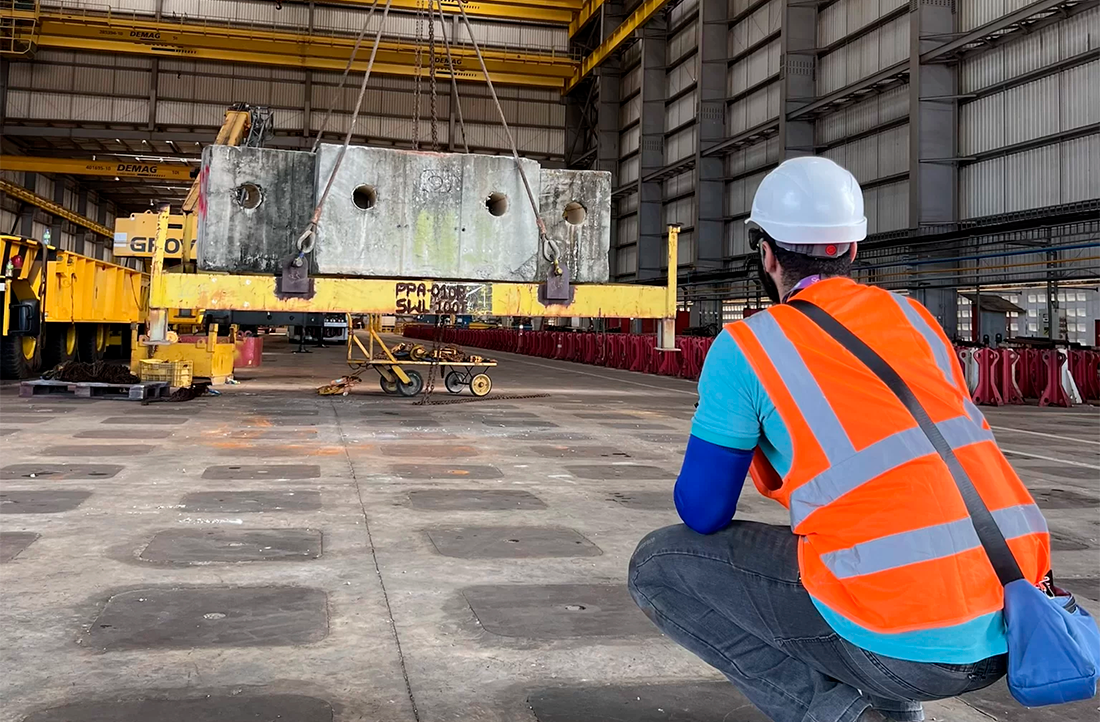 Pessoa agachada trajando colete de segurança laranja e capacete observando teste de içamento de carga.