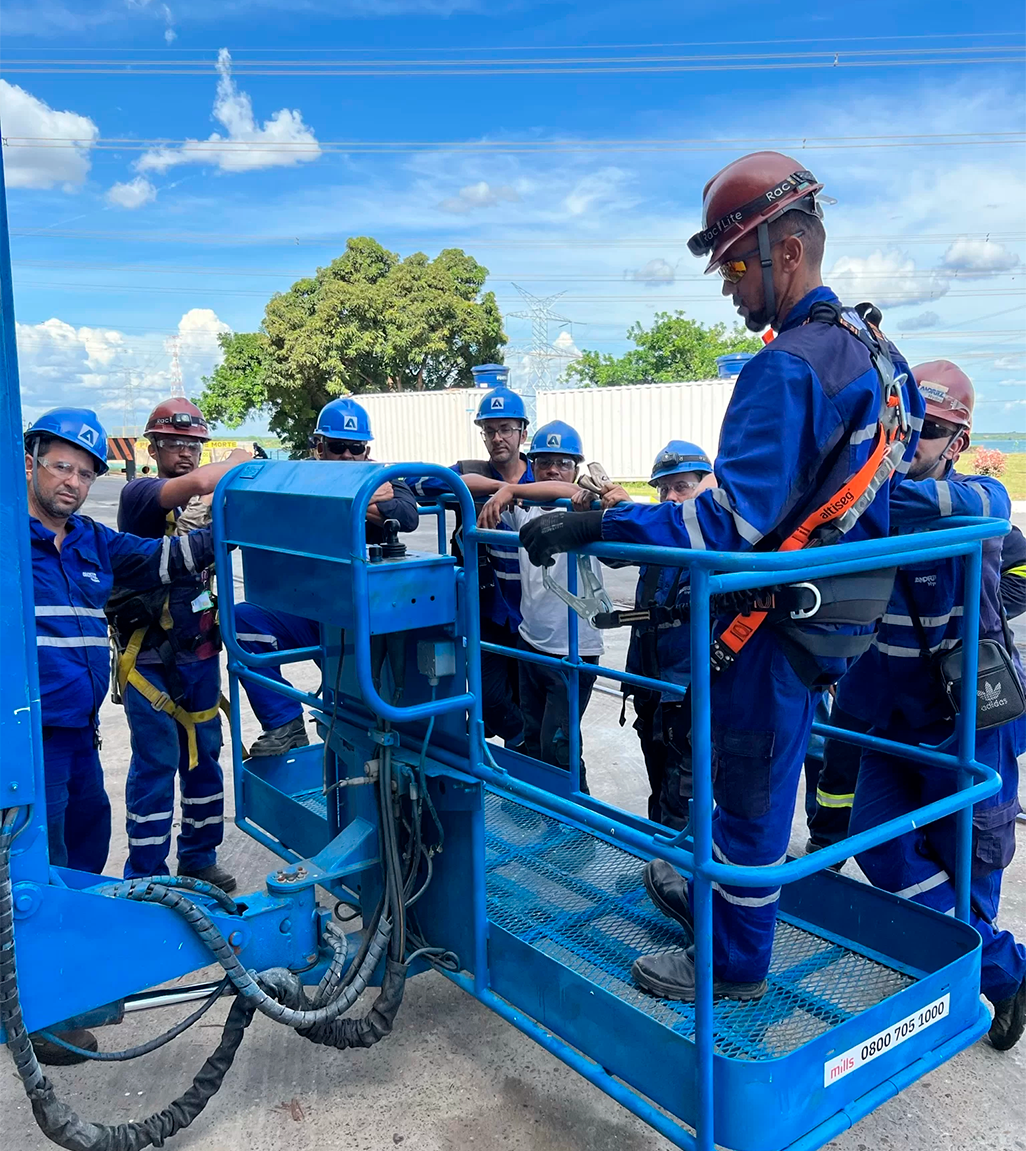 Pessoas trajando capacetes, macacão, óculos de proteção e arnês ao redor de um homem trajado da mesma forma que está dentro de um elevador de obra e conectado a ele.